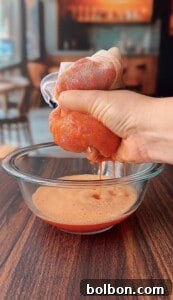 A hand squeezes a mesh bag filled with blended tomatoes over a glass bowl, extracting tomato juice onto a wooden table in a cozy kitchen setting.