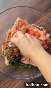 A hand mixing ground meat with chopped herbs, spices, and red seasoning in a glass bowl on a wooden surface.
