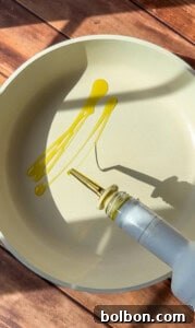 A stream of olive oil is being poured from a bottle into a light-colored frying pan, ready to cook the viral blanket dumplings, while sunlight casts shadows on the wooden surface.