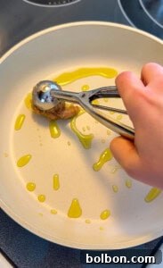 A hand uses a metal ice cream scoop to place a portion of Viral Blanket Dumplings onto a white frying pan drizzled with olive oil. The pan sits on a stovetop, ready to cook these trending treats.