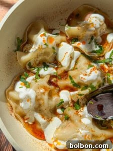 A close-up of Viral Blanket Dumplings topped with dollops of yogurt, a drizzle of red chili oil, chopped fresh herbs, and a sprinkle of spices, served in a bowl with a wooden spoon.