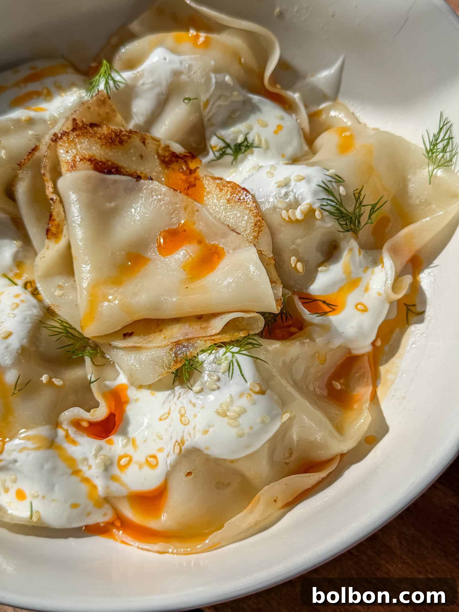 A close-up of Viral Blanket Dumplings topped with creamy white sauce, orange chili oil, sesame seeds, and fresh dill, served in a white bowl.