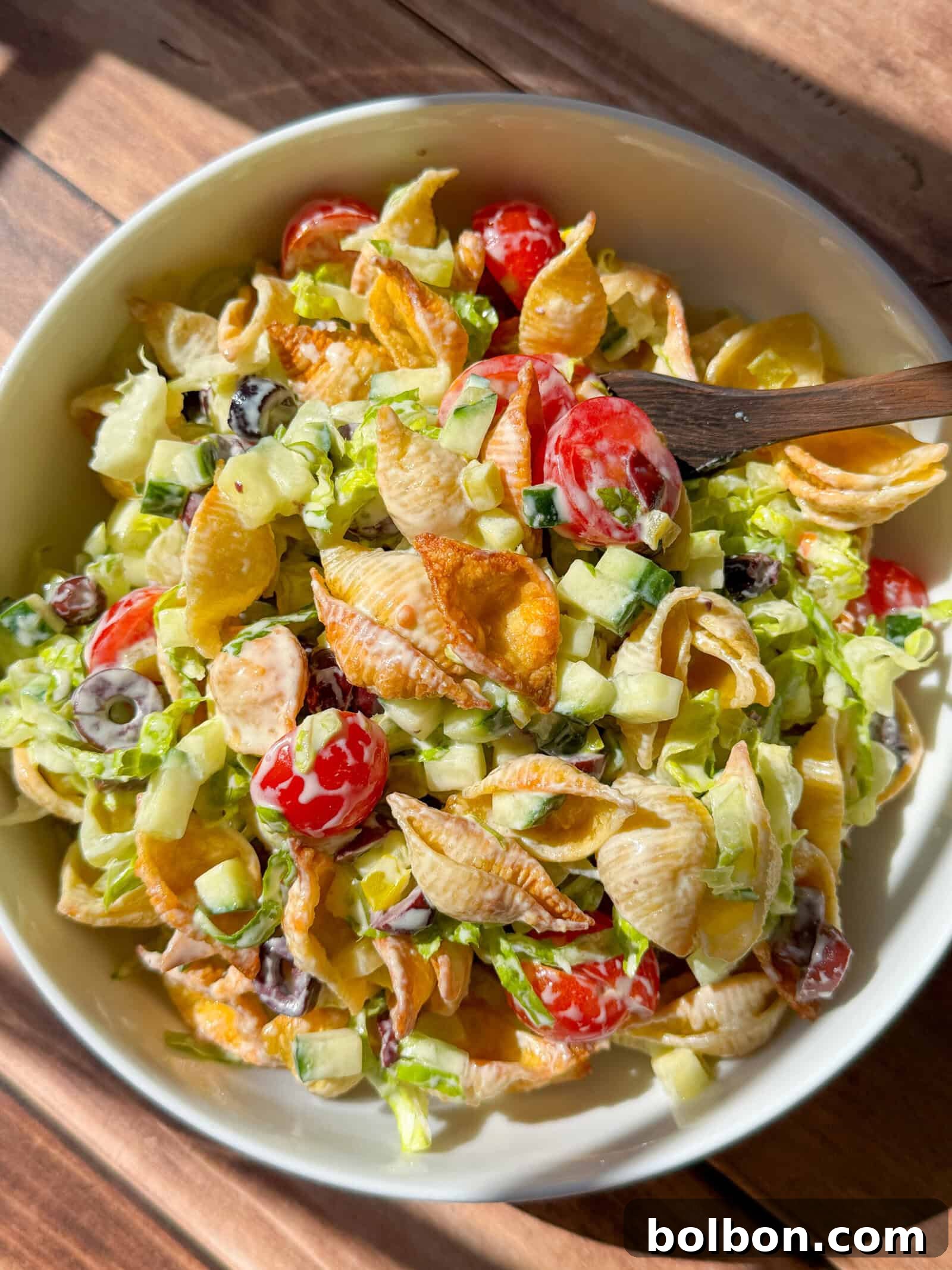 A vibrant bowl of crispy pasta salad featuring golden shell pasta, juicy cherry tomatoes, sliced black olives, crisp chopped lettuce, fresh cucumbers, and a rich, creamy dressing. A wooden spoon rests invitingly in the bowl, illuminated by natural sunlight.