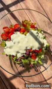 A glass bowl filled with chopped romaine lettuce, cherry tomatoes, sliced black olives, diced green peppers, and topped with a generous amount of creamy salad dressing, sitting on a wooden surface in sunlight.