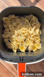 Uncooked pasta shells topped with shredded cheese and breadcrumbs in an air fryer basket, placed on a wooden surface with sunlight casting shadows.
