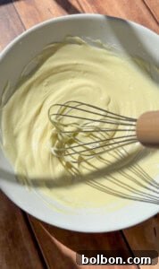 A white bowl filled with creamy yellow batter is being mixed with a metal whisk with a wooden handle, placed on a wooden surface in bright sunlight.
