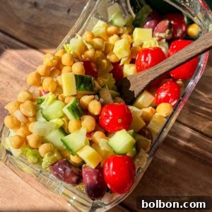 A glass container tipped on its side spills a colorful salad with chickpeas, grape tomatoes, cucumber, cheese cubes, and olives onto a wooden surface. A wooden fork rests inside the container.