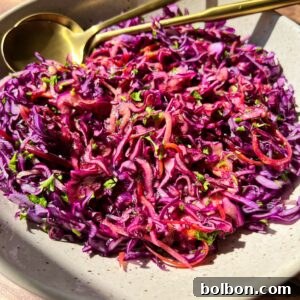 A bowl of vibrant red cabbage slaw with finely shredded purple cabbage, carrots, and herbs, topped with a light vinaigrette. A gold fork and spoon rest beside the dish on a wooden table.