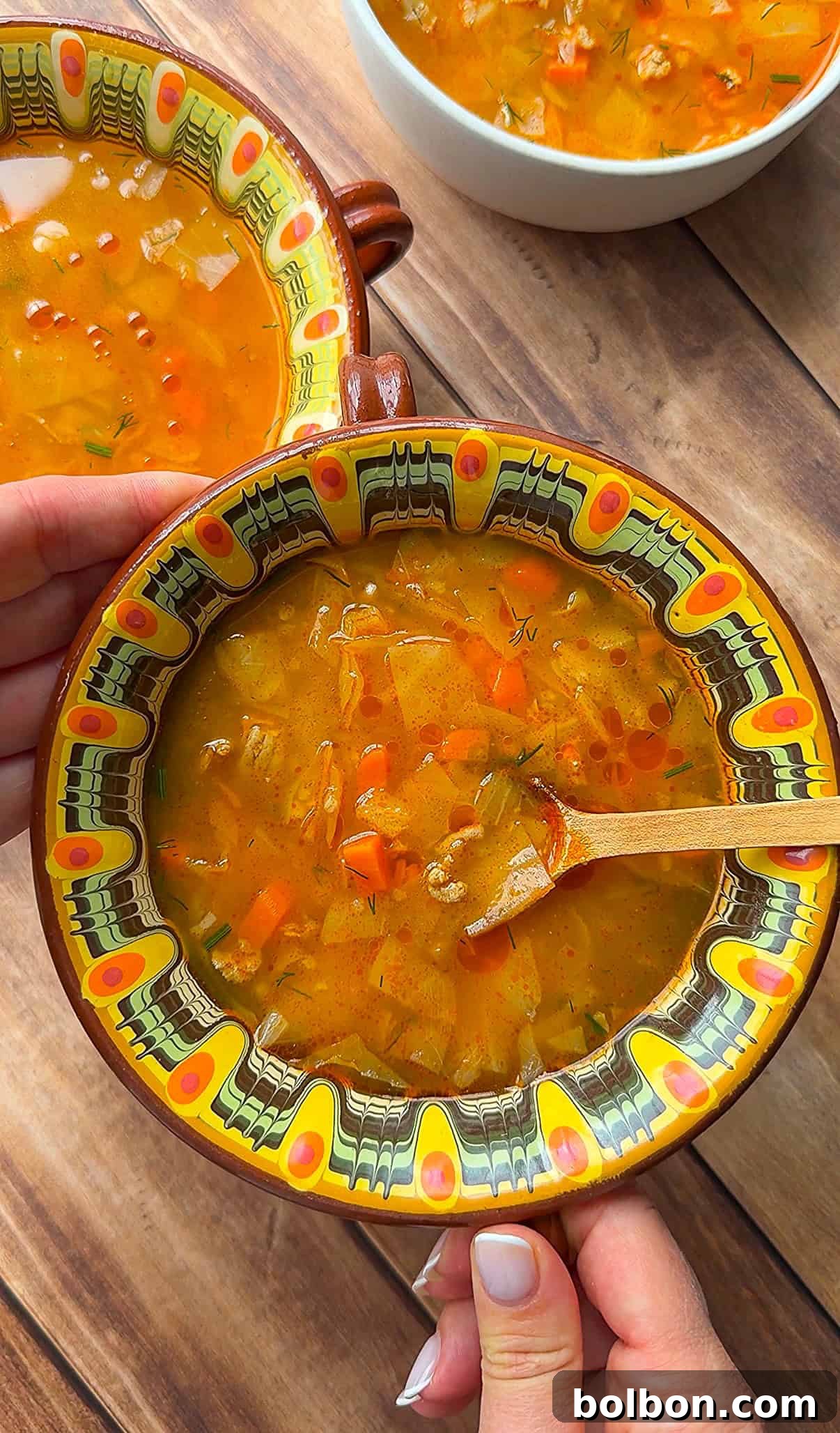 A hand holds a vibrant, patterned bowl overflowing with vegetable soup, enriched with lean ground turkey and tender cabbage. Another hand gently stirs the soup with a wooden spoon, while two more steaming bowls sit invitingly on a rustic wooden table in the soft background. The scene evokes comfort, warmth, and wholesome nourishment.