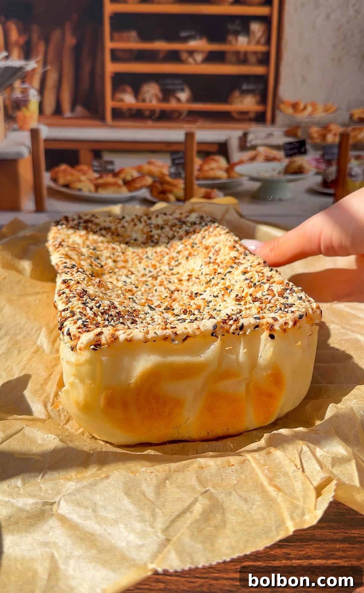 A hand holds a rectangular, seeded bread—reminiscent of the viral Blender Rice Bread—covered in sesame and poppy seeds, resting on parchment paper with a bakery display of breads and pastries in the background.