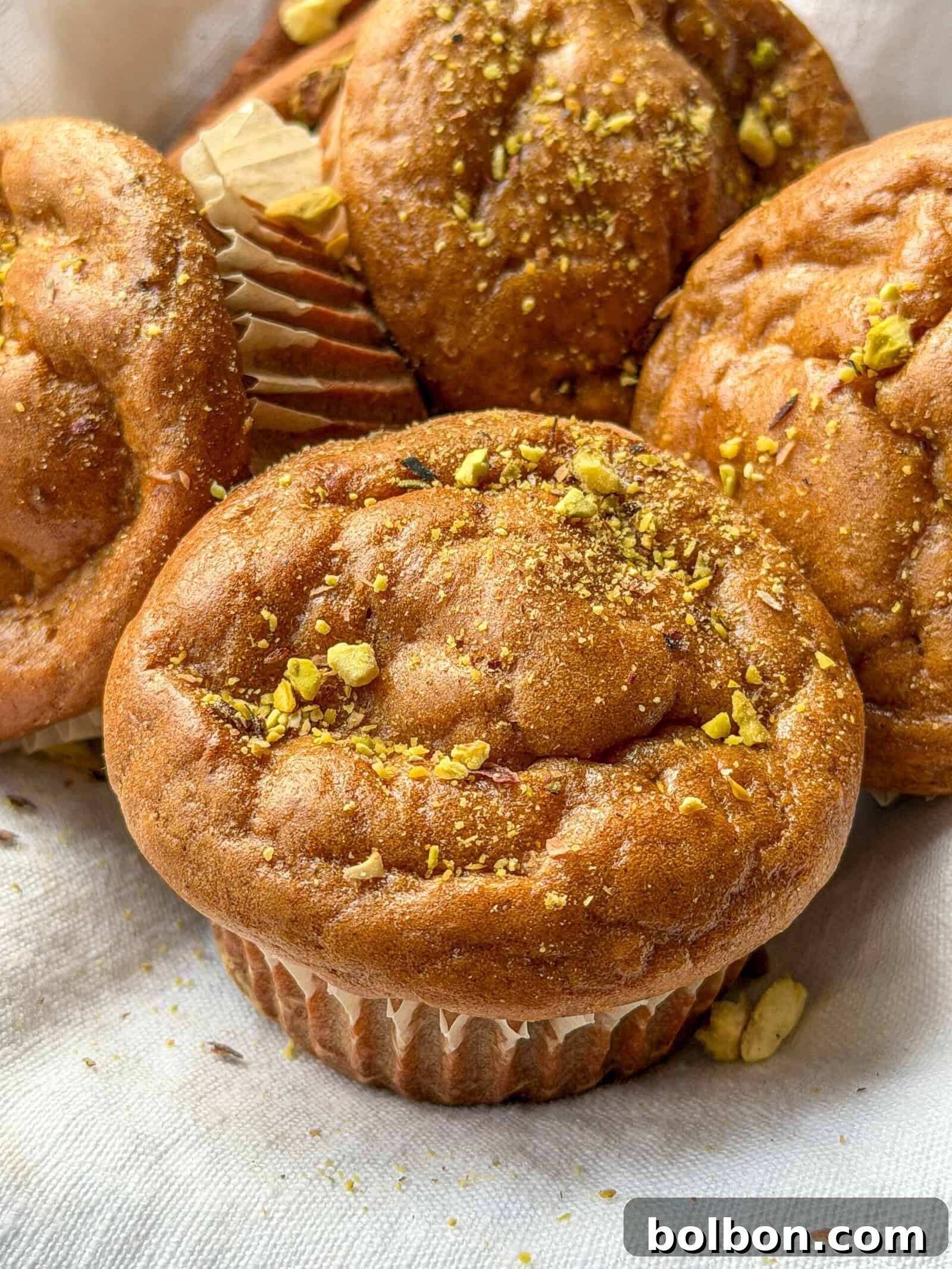 Wholesome Sweet Potato Muffins 2 A close-up of healthy sweet potato muffins topped with crushed pistachios, resting on a light-colored cloth. The muffins appear fluffy and moist, with slightly cracked tops and paper liners, ready to be enjoyed as a wholesome snack.