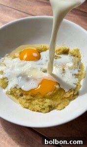 Milk being poured into a white bowl containing two raw eggs and mashed bananas, perfect for preparing Healthy Sweet Potato Muffins, set on a rustic wooden surface, highlighting the liquid ingredients.
