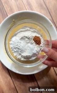A hand holds a glass bowl containing flour, baking powder, and cinnamon above a larger white bowl with a yellow mixture on a wooden surface—perfect for making Healthy Sweet Potato Muffins, demonstrating the dry ingredient addition.