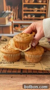 A hand lifts one of three muffins from a cooling rack lined with bakery-themed paper, with shelves of bread and baked goods visible in the background, signaling fresh baked goodness.