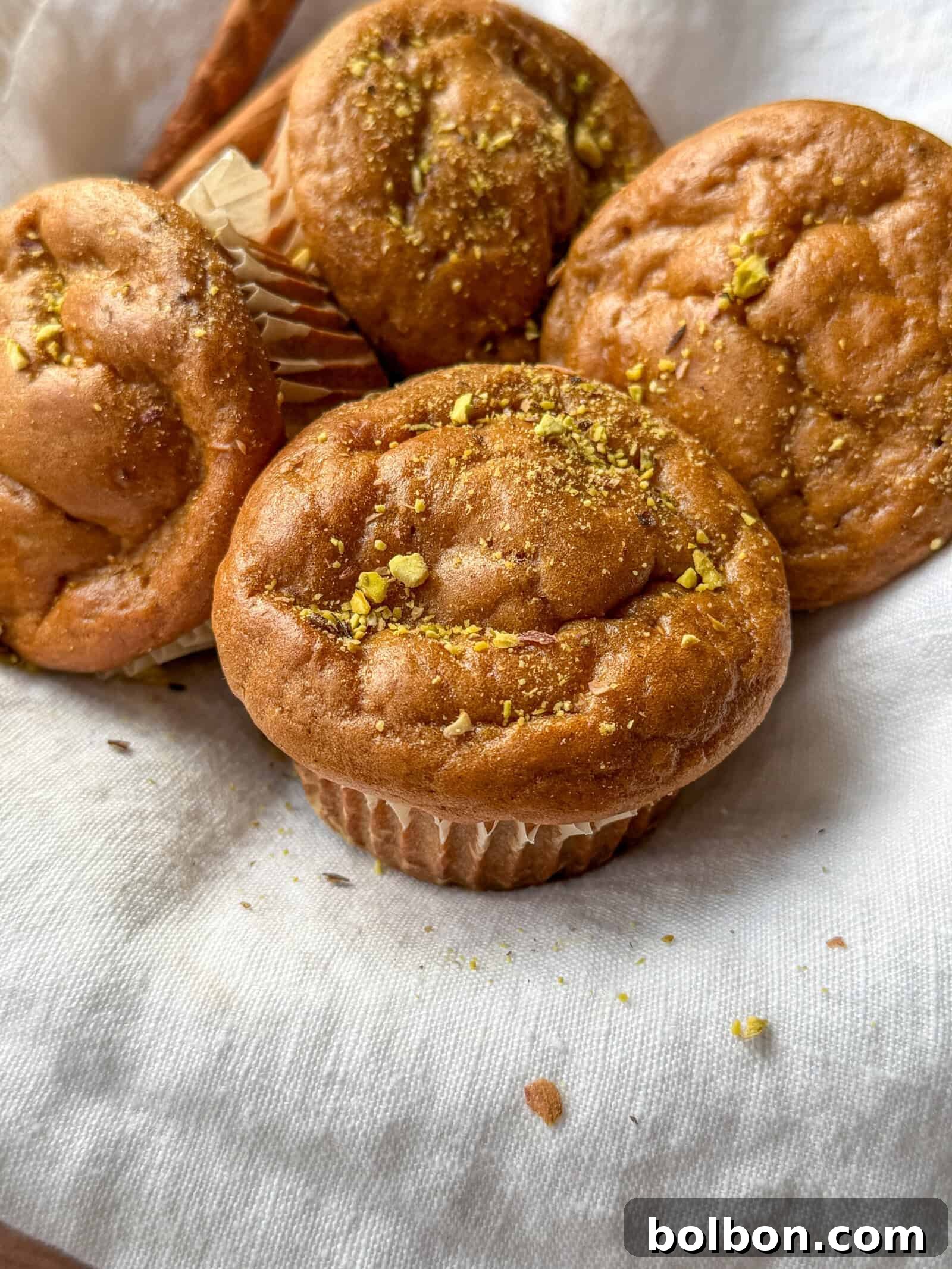 Wholesome Sweet Potato Muffins 3 Four golden brown muffins sprinkled with crushed pistachios rest on a white cloth, with one muffin slightly separated in front, showcasing their soft and fluffy texture, inviting a taste.