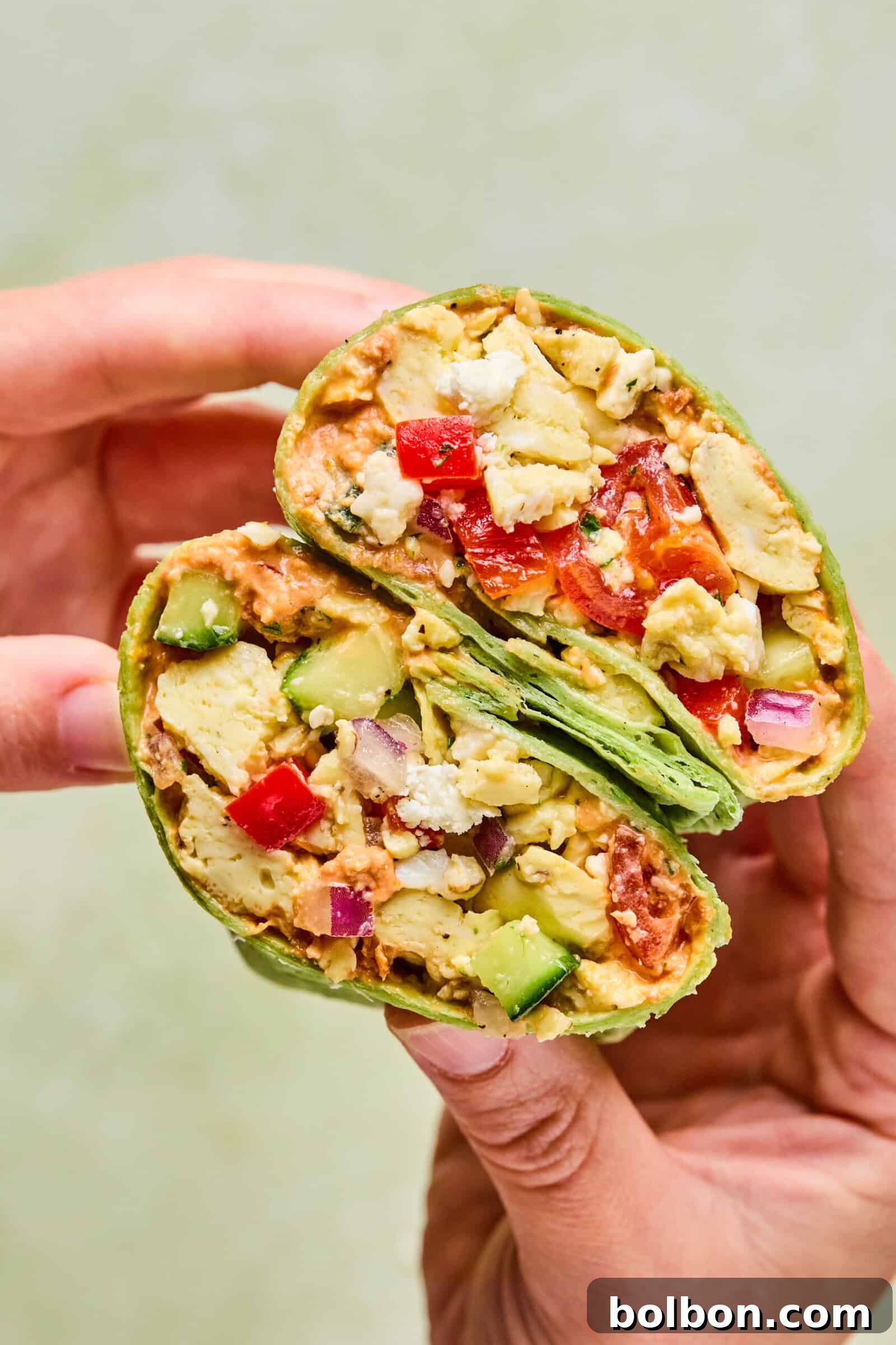 A hand holds a halved Mediterranean Breakfast Wrap filled with scrambled tofu, diced red and green bell peppers, onions, avocado, and other vegetables in a green spinach tortilla.