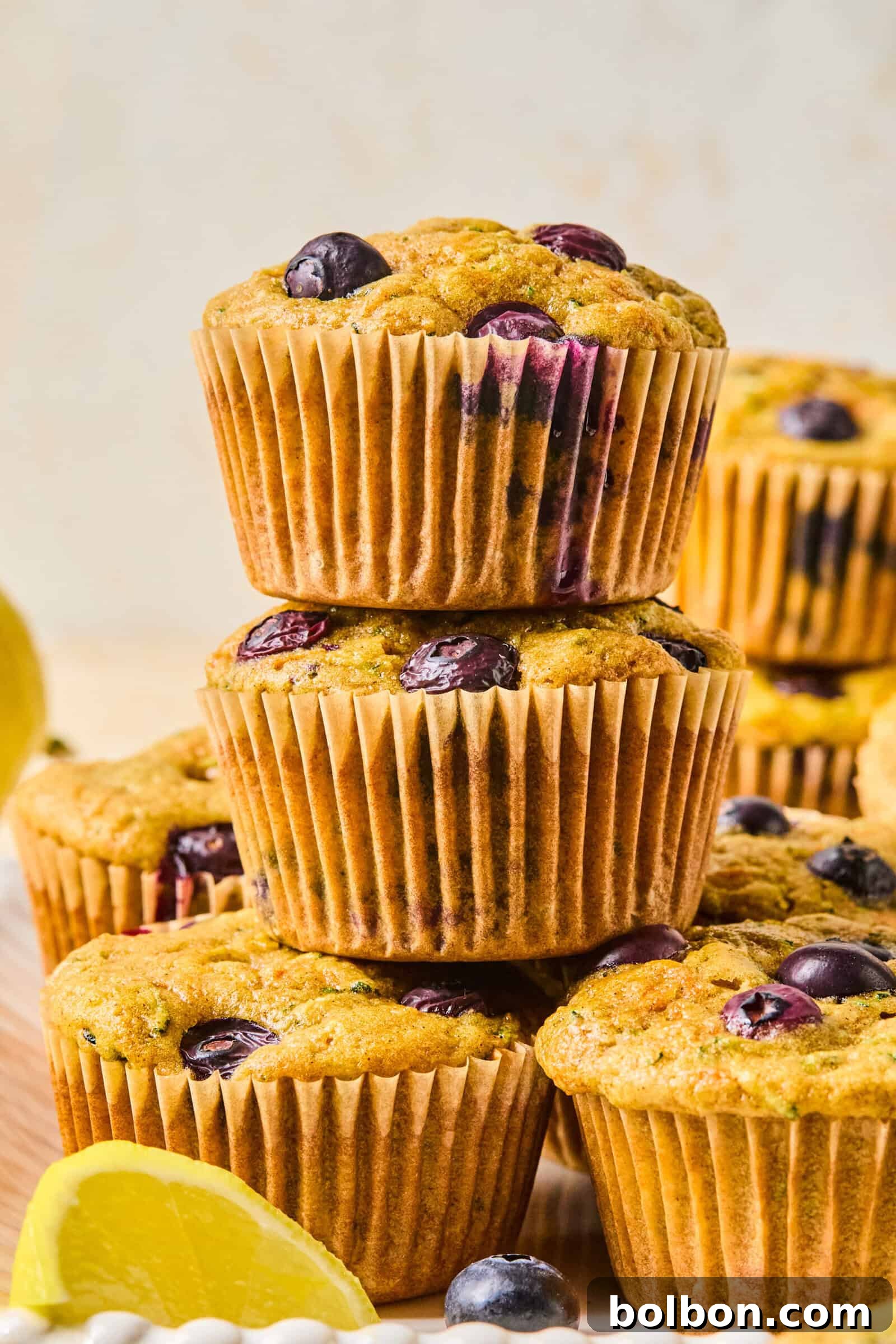 Garden Burst Lemon Blueberry Muffins 2 A stack of three golden-brown lemon blueberry veggie muffins, topped with a sprinkle of oats, sits invitingly on a rustic wooden surface. Several more muffins are blurred in the background, alongside a fresh lemon wedge, evoking a cozy, wholesome morning scene. These muffins are perfect to accompany a savory Mediterranean Breakfast Wrap for a truly delightful and wholesome start to your day.