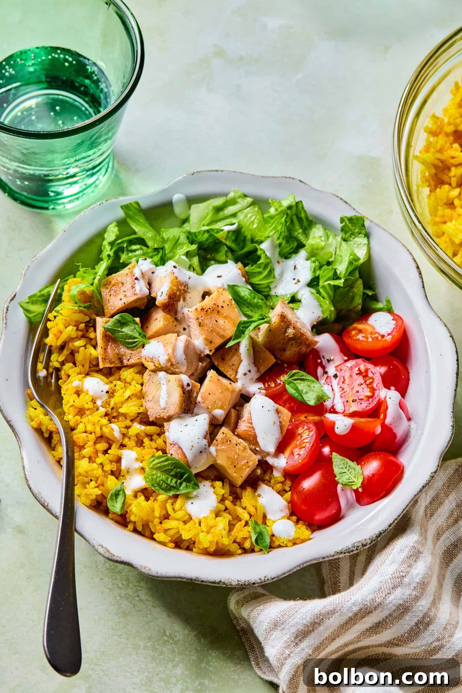 A close-up shot of the chicken rice bowl, showing the texture of the yellow rice, the diced chicken, fresh tomatoes, and herbs, all drizzled with a creamy white sauce.