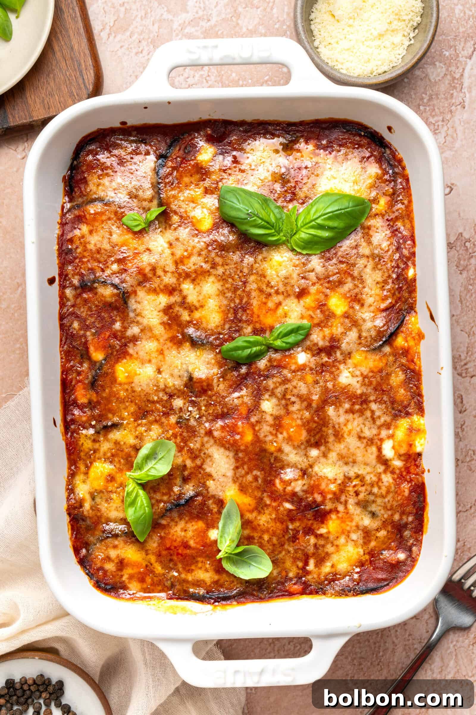 Classic Eggplant Parmesan (Parmigiana di Melanzane) Recipe 2 A baked eggplant parmesan casserole in a white dish, topped with melted cheese, tomato sauce, and fresh basil leaves, with a bowl of grated cheese nearby.