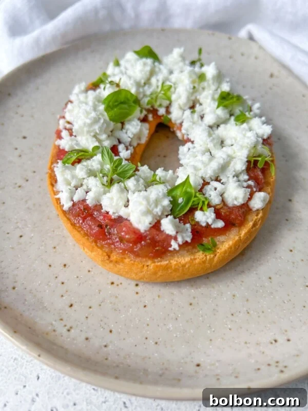 Pan con Tomate: Authentic Spanish Tomato Bread Recipe & How to Make It 4 A toasted bagel half topped with tomato sauce, crumbled white cheese, and fresh basil leaves sits on a speckled beige plate—an irresistible twist on Greek Bruschetta.