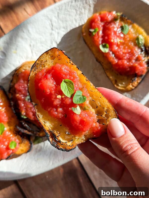 Pan con Tomate: Authentic Spanish Tomato Bread Recipe & How to Make It 7 A hand holds a slice of toasted bread topped with crushed tomatoes and fresh green herbs, reminiscent of flavors from an Italian eggplant parmesan recipe, with more slices on a white plate in the background on a sunlit wooden surface.