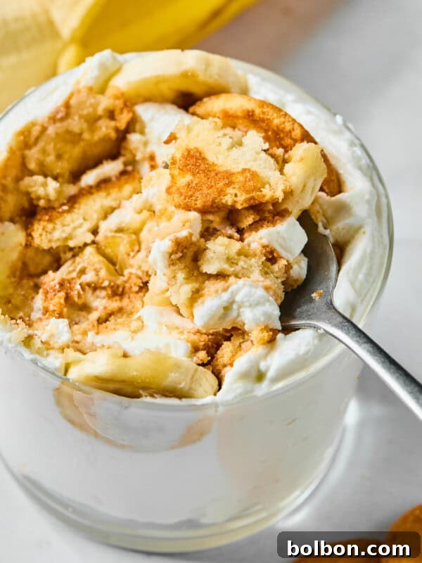 A close-up of a glass filled with banana pudding, whipped cream, banana slices, and crumbled vanilla wafers, with a spoon scooping out a portion. A banana and wafers are visible in the background.