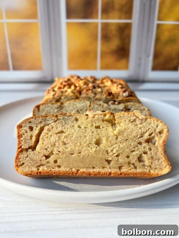 A sliced loaf of moist blender banana bread on a white plate, displayed in front of a window with autumn foliage visible outside. The bread has a golden crust and is topped with chopped nuts.