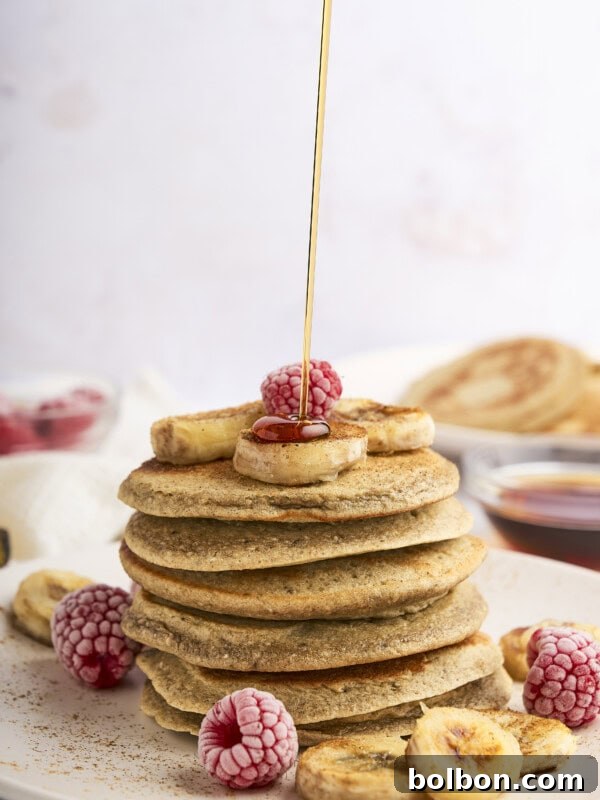 A stack of pancakes topped with banana slices and raspberries on a white plate. Syrup is being poured over the top. In the background, there are more pancakes and a small bowl of syrup.