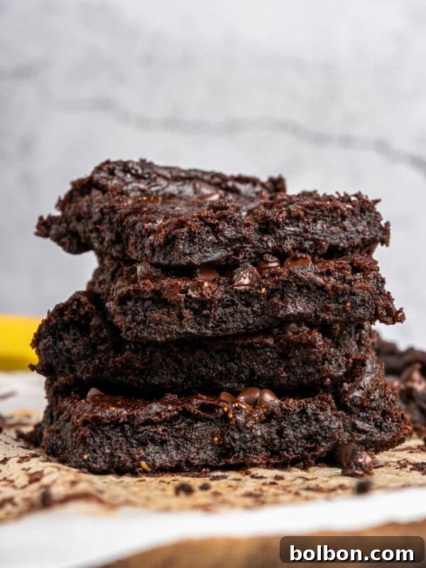 A close-up of a stack of three gooey chocolate brownies on parchment paper. The brownies are rich and moist, with visible chocolate chips embedded in the batter. The background is a simple, textured surface.