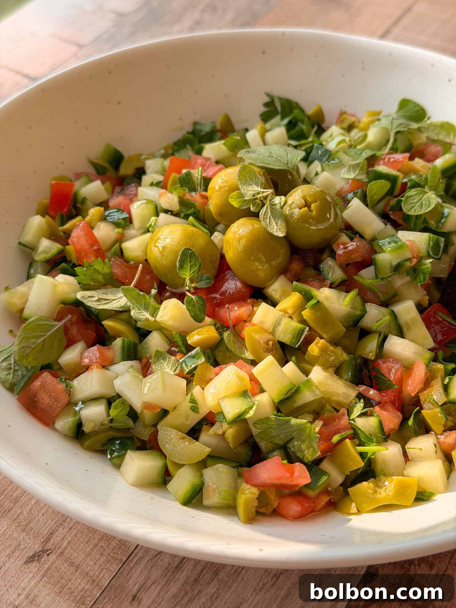 Fresh & Easy Mediterranean Bread Dipping Oil Recipe 2 A bowl of chopped salad with cucumbers, tomatoes, green olives, pickles, and fresh herbs is served in a white dish on a wooden surface—perfect alongside your favorite bread dipping oil recipe.