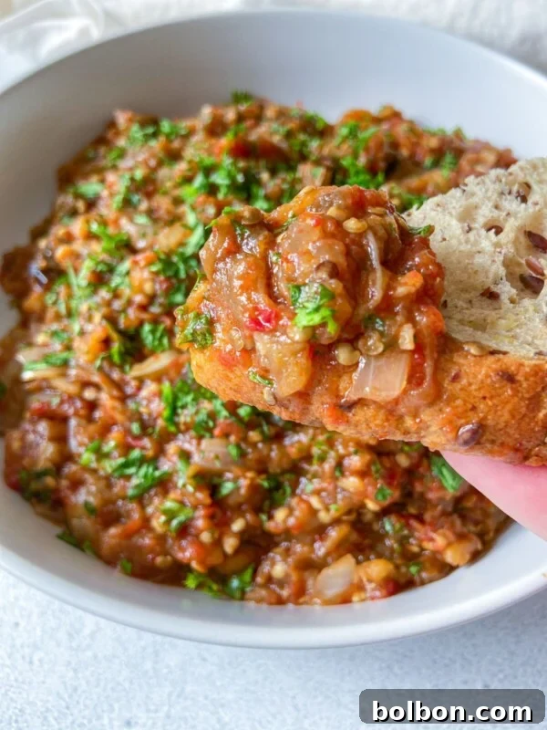 Fresh & Easy Mediterranean Bread Dipping Oil Recipe 7 A close-up of a piece of bread topped with a chunky eggplant dip, garnished with chopped fresh herbs, held above a bowl filled with more of the same dip.