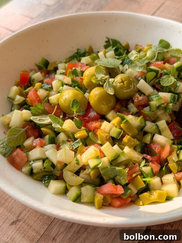 Fresh & Easy Mediterranean Bread Dipping Oil Recipe 8 A bowl of chopped salad with cucumbers, tomatoes, green olives, pickles, and fresh herbs is served in a white dish on a wooden surface—perfect alongside your favorite bread dipping oil recipe.
