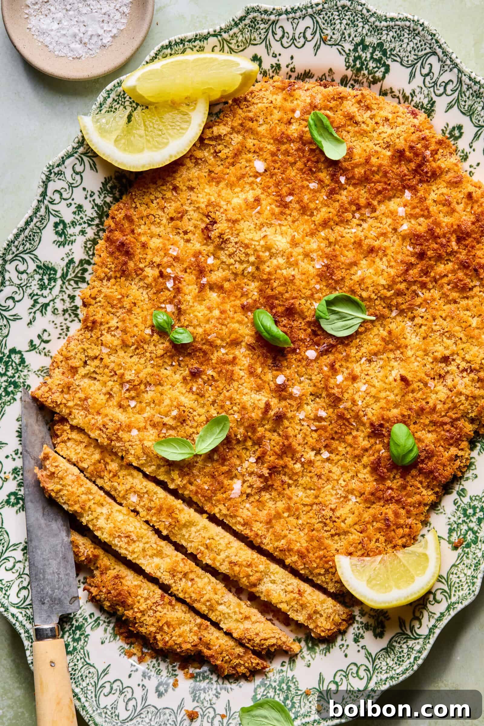 A large, breaded and baked schnitzel on a decorative plate, garnished with fresh basil leaves and lemon wedges, with a knife and several sliced pieces at one end. A small bowl of salt is in the background.