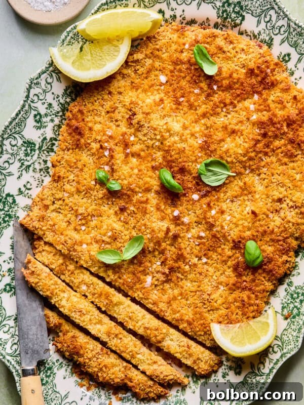 A large, breaded and baked schnitzel on a decorative plate, garnished with fresh basil leaves and lemon wedges, with a knife and several sliced pieces at one end. A small bowl of salt is in the background.