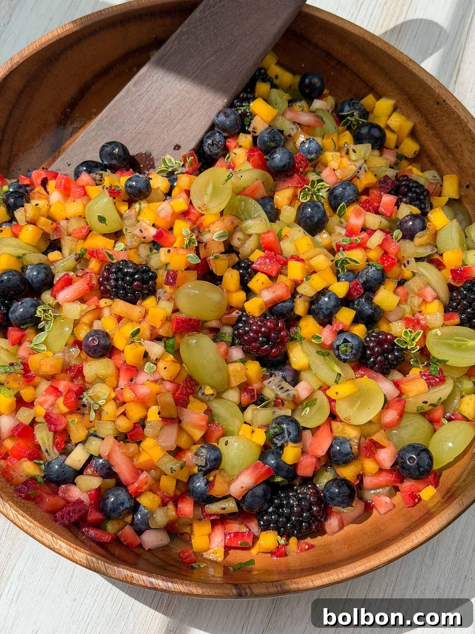 A wooden bowl filled with a colorful fruit salad featuring diced mango, watermelon, strawberries, green grapes, blueberries, blackberries, and a few small green herbs, with a wooden serving spoon.