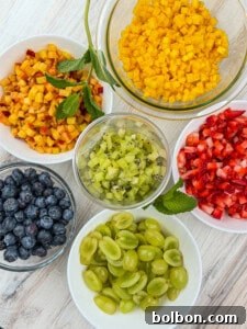 Six bowls of chopped fruit on a light wooden surface, including diced mango, diced peaches, chopped strawberries, blueberries, sliced green grapes, and diced kiwi, with mint leaves for garnish.
