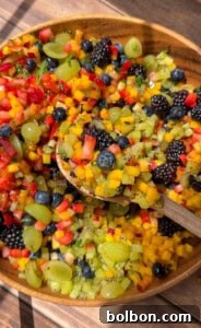 A wooden bowl filled with a colorful fruit salad, including diced mango, kiwi, strawberries, grapes, blueberries, and blackberries. A wooden spoon scoops up a portion of the vibrant mix.