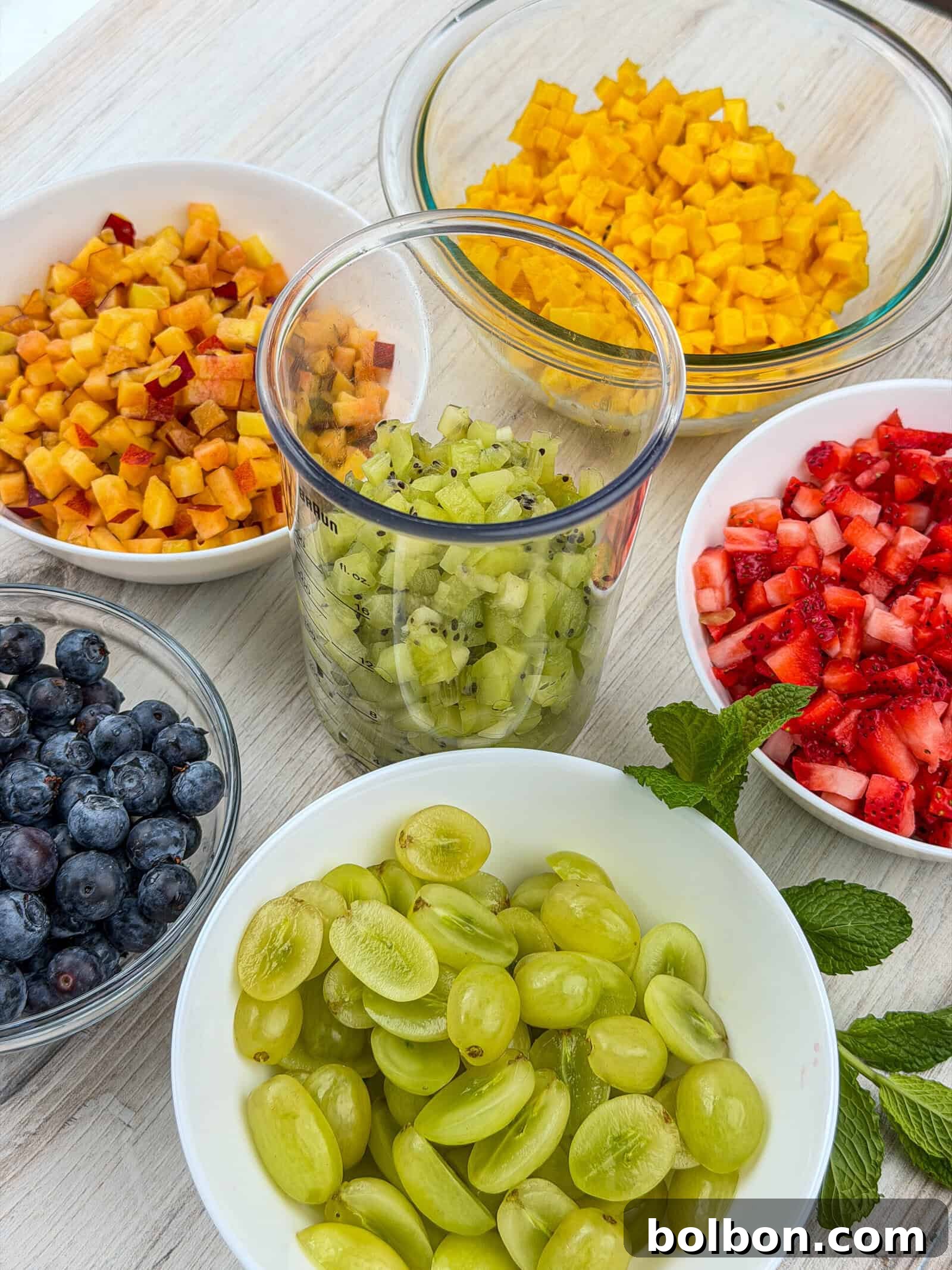 Bowls of chopped fruit on a light surface, including blueberries, green grapes, strawberries, kiwi, peach, and mango. There are also a couple of sprigs of fresh mint near the bowls.