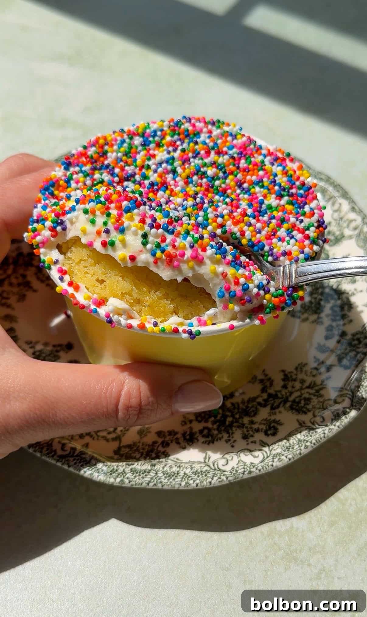A hand holds a yellow cup filled with Viral Dot Cake, topped with white frosting and colorful rainbow sprinkles. A spoon scoops out a bite, revealing the soft yellow cake inside. The cup sits on a decorative floral-patterned saucer.