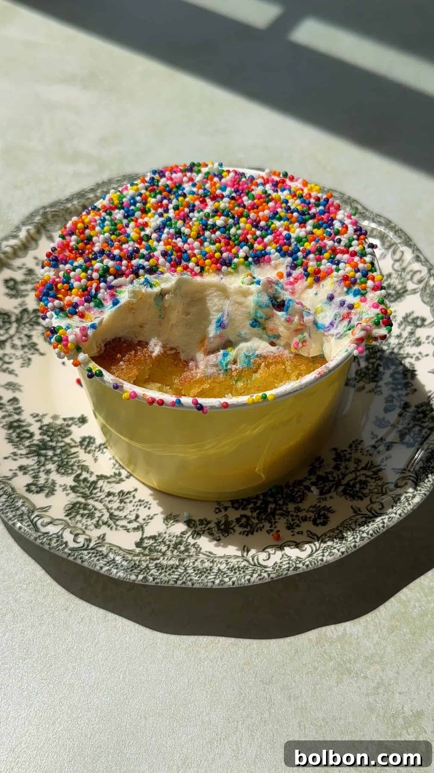 A small round Viral Dot Cake in a yellow cup, topped with white frosting and colorful sprinkles, sits on a patterned plate. A portion has been eaten, revealing the cake and frosting layers inside. Sunlight casts shadows on the table.