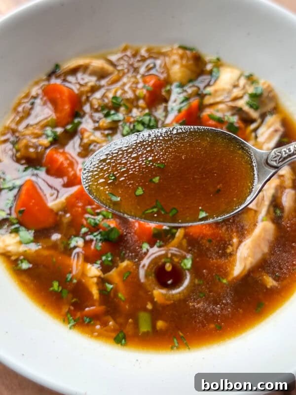Viral No-Water Chicken Soup Recipe That Uses Just Broth 7 A close-up of No Water Chicken Soup with a spoonful of clear brown broth held above it, showing pieces of chicken, chopped carrots, and fresh herbs on top—perfect for a rich, waterless chicken soup recipe.