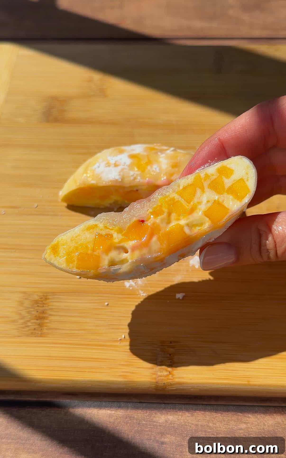 Viral Strawberry & Mango Rice Paper Mochi Recipe 3 A hand holds half of a rice paper mochi dessert filled with creamy white and orange mango pieces on a wooden cutting board, with sunlight casting soft shadows.