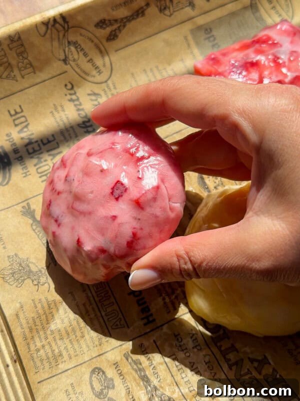 Viral Strawberry & Mango Rice Paper Mochi Recipe 8 A hand holding a round, pink, textured dessert with red specks rests on brown printed parchment paper in bright sunlight—reminiscent of the vibrant hues in a No Water Chicken Soup recipe. Another yellow item is partially visible nearby.