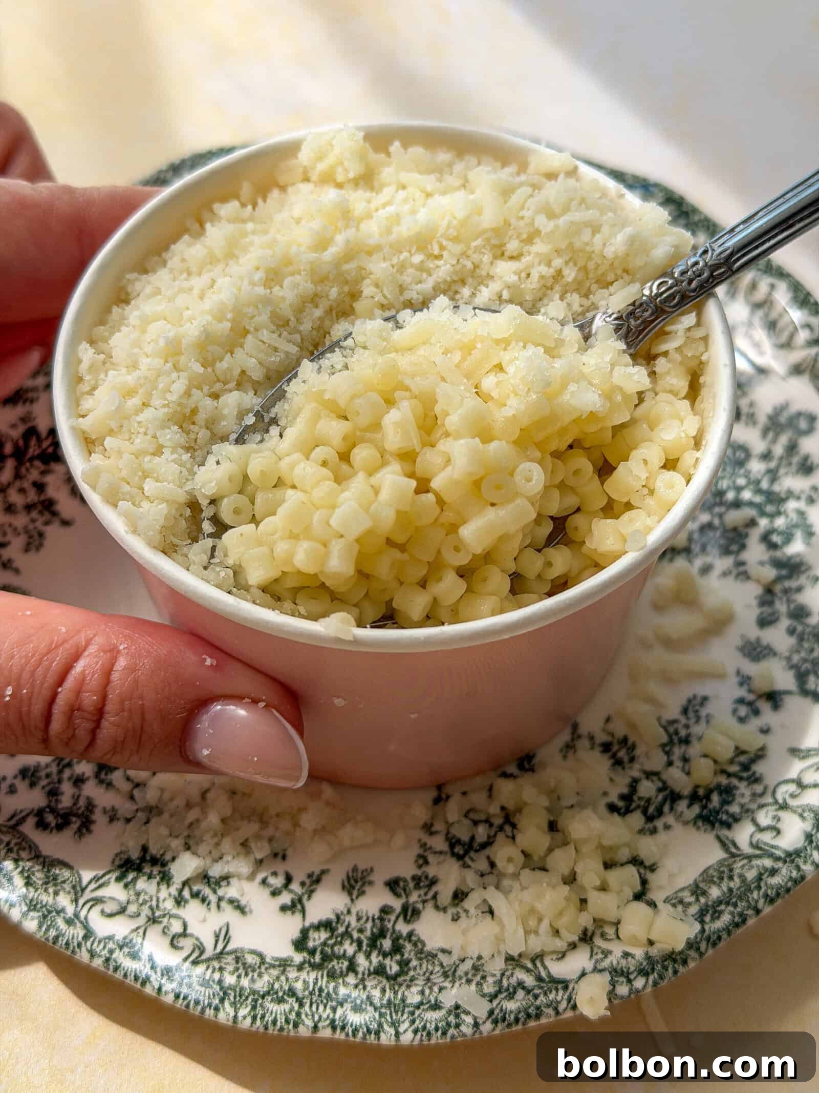 A hand holds a small cup filled with tiny pasta and topped with grated cheese. A spoon is scooping some pasta, and the cup rests on a decorative green and white plate, with cheese scattered around.