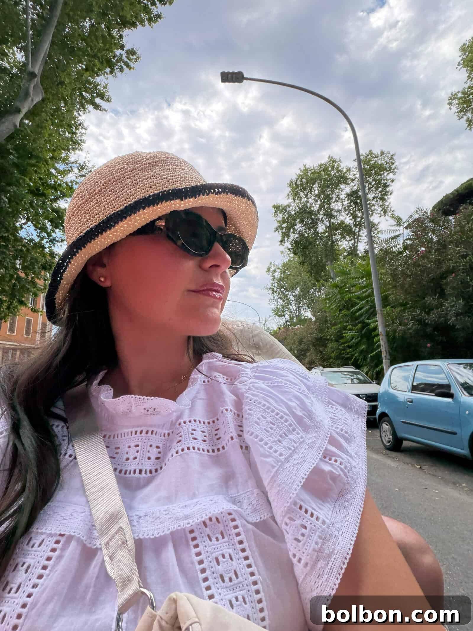 Woman wearing a straw hat, sunglasses, and a white embroidered blouse sits outdoors on a street lined with trees and parked cars, looking off to the side under a partly cloudy sky.