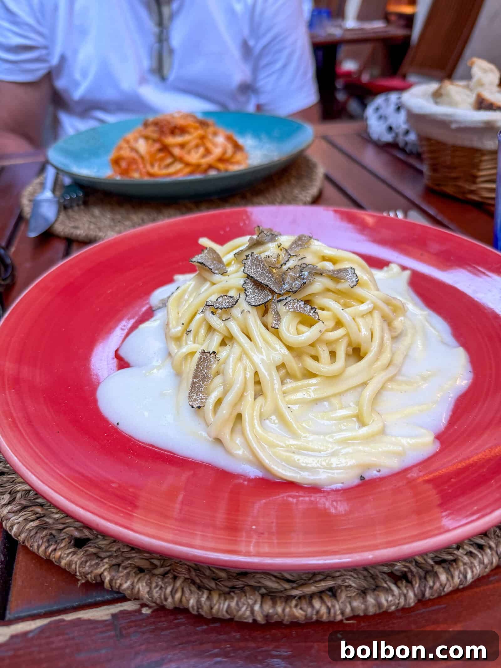 Close-up of a plate of pasta with creamy white sauce and truffle slices on a red plate. In the background, another plate of pasta with red sauce and a partially visible person sitting at a wooden table.