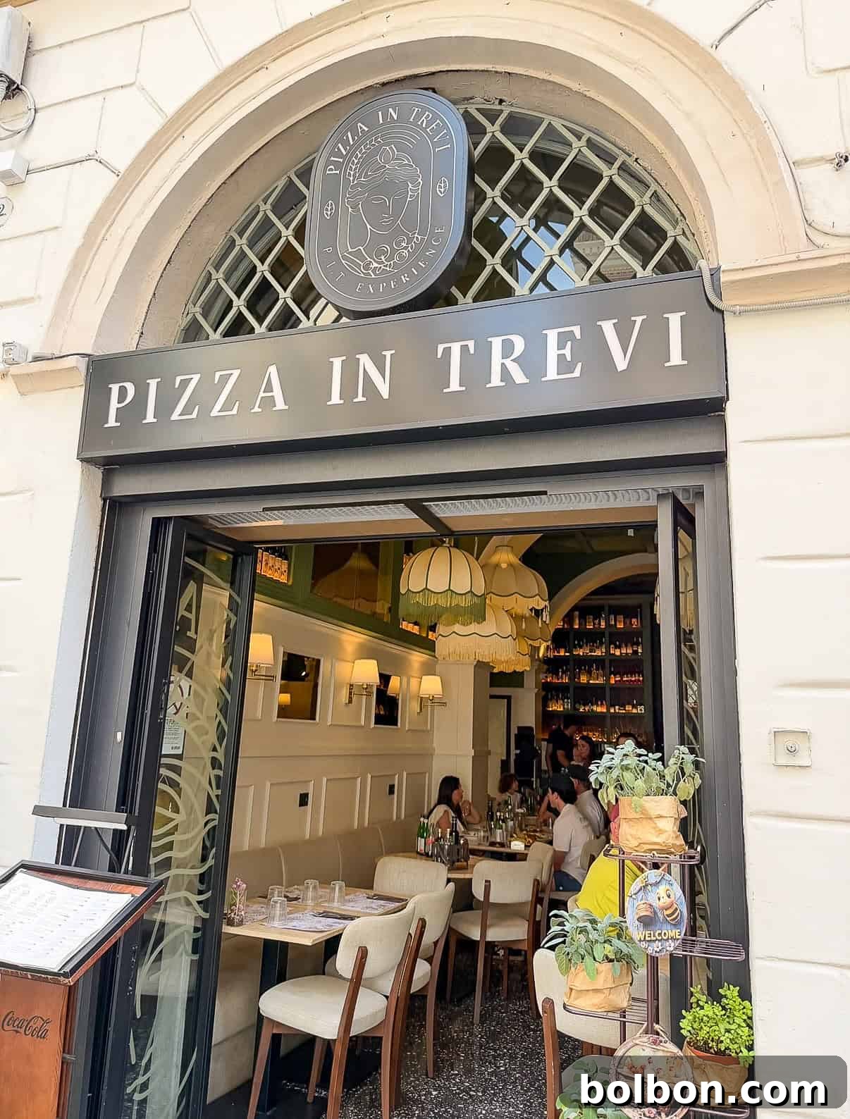 Street view of "Pizza in Trevi" restaurant with an arched entrance, open doors, visible dining tables, chandeliers, and people sitting inside. A menu stand and a plant display are at the entrance.