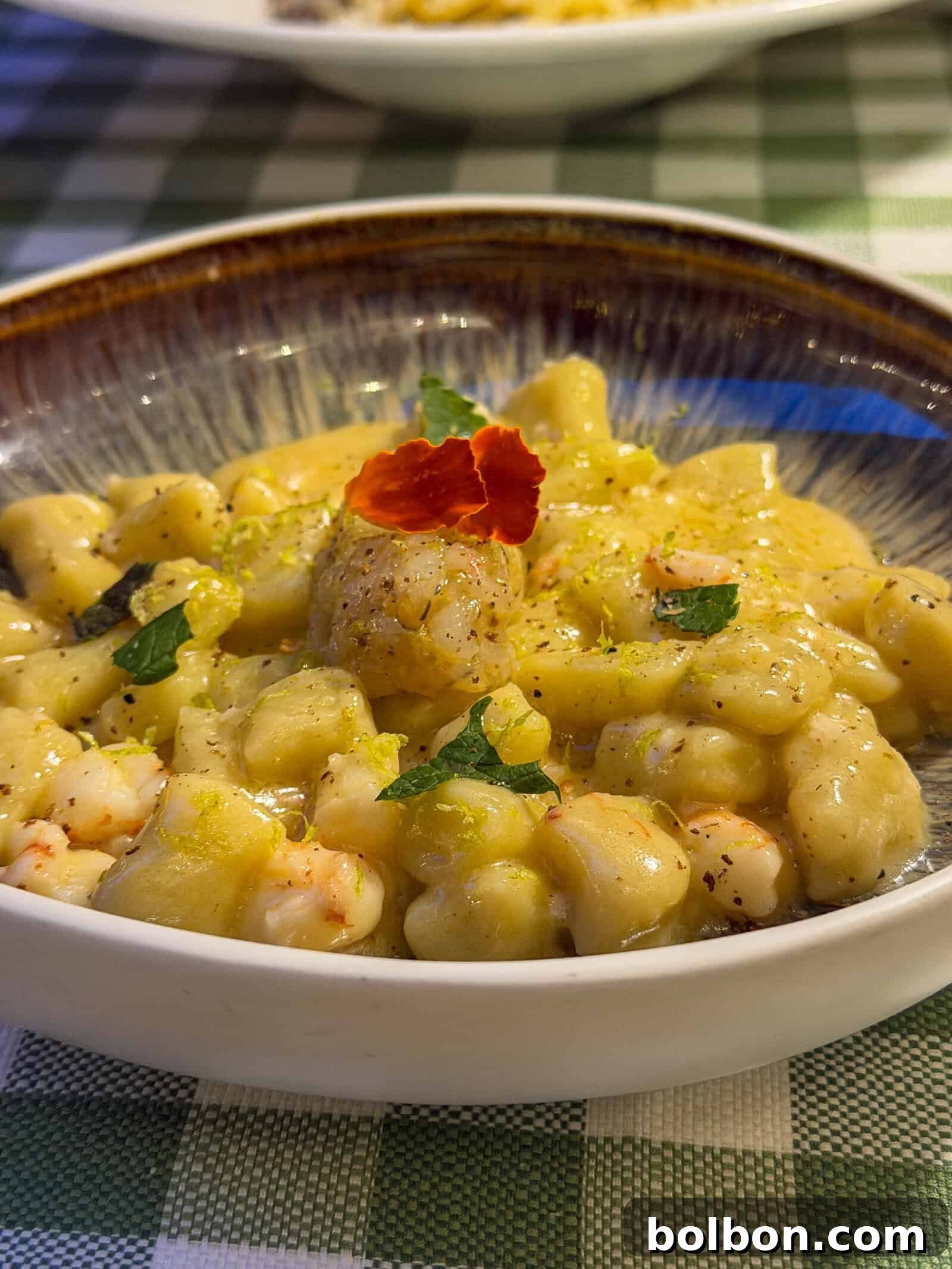 A close-up of a bowl of gnocchi in a creamy yellow sauce, garnished with herbs and an edible red flower, served on a green and white checkered tablecloth.