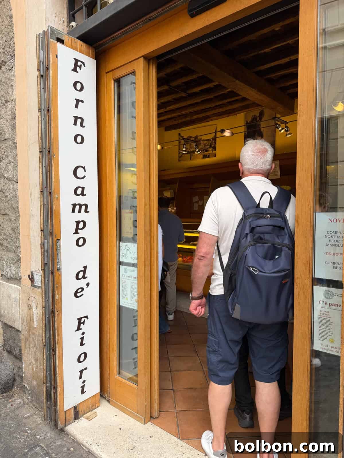 A man with a backpack enters Forno Campo de’ Fiori, a bakery with a white sign and wooden door, as people stand inside near the counter.