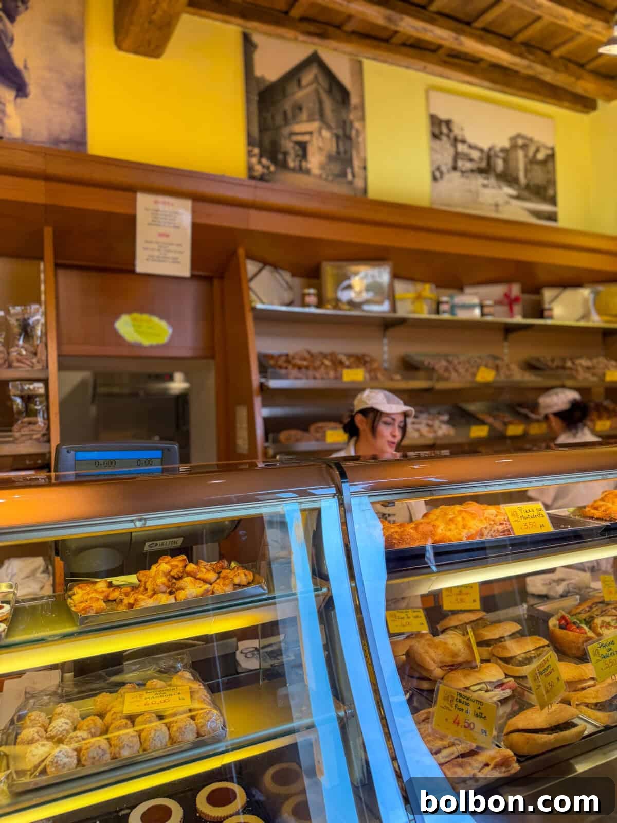 A bakery with glass display cases filled with various pastries and breads. A worker wearing a white cap stands behind the counter. Yellow price tags are visible, and shelves with more baked goods and vintage photos are on the wall.